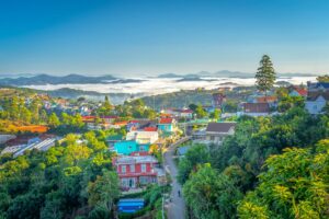Elevated view over Dalat town with colorful houses set among pine forest and greenhouses, morning mist rolling across the valley toward distant mountains