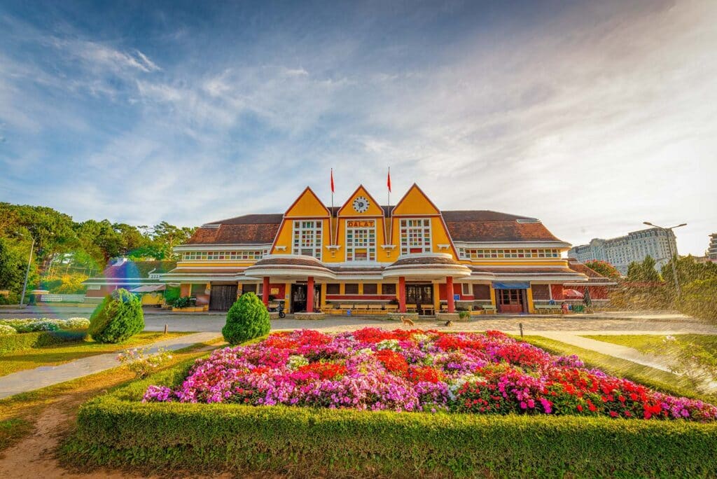 The yellow Art Deco facade of Dalat railway station with its three pointed roofs and DALAT sign, fronted by a colorful flower garden under a blue sky, one of the classic things to do in Dalat