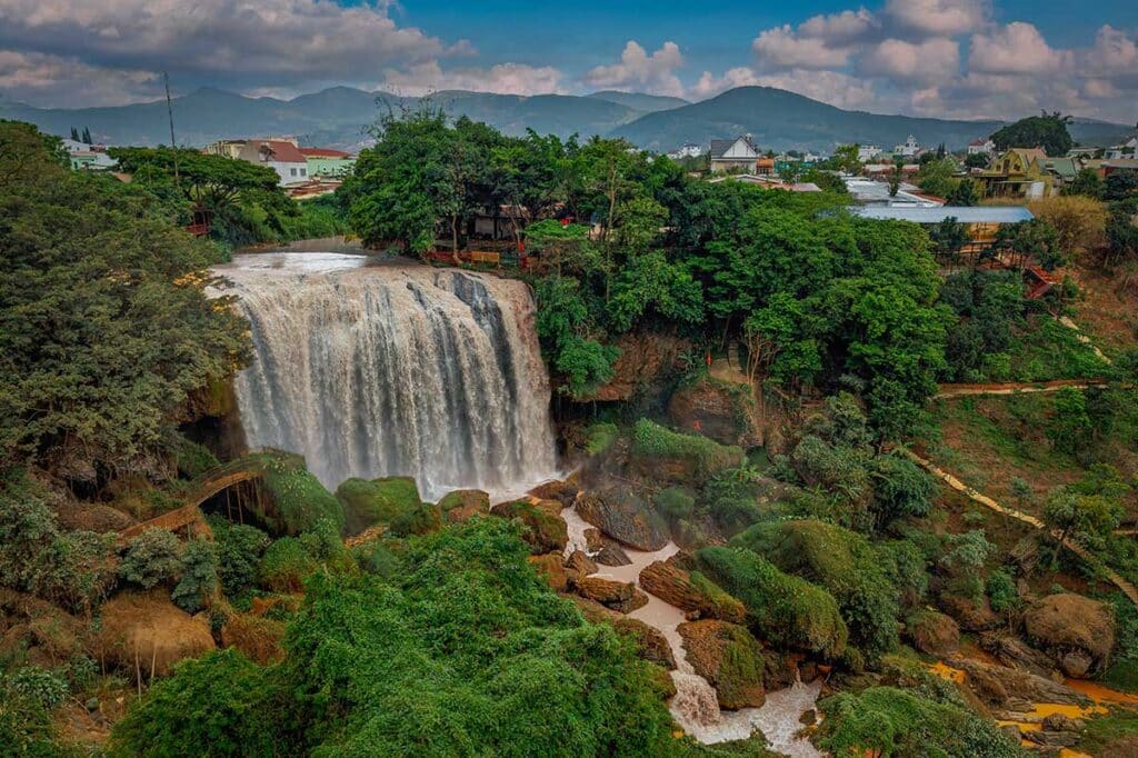 Elephant Waterfall in Dalat crashing down a broad rock face into a churning pool, surrounded by moss-covered boulders and green forest, with houses and mountains behind