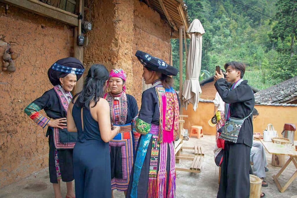 Visitors talking with Hmong and Dao women in traditional dress in the courtyard of a clay house in a village on the Dong Van Karst Plateau