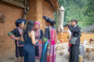 Visitors talking with Hmong and Dao women in traditional dress in the courtyard of a clay house in a village on the Dong Van Karst Plateau