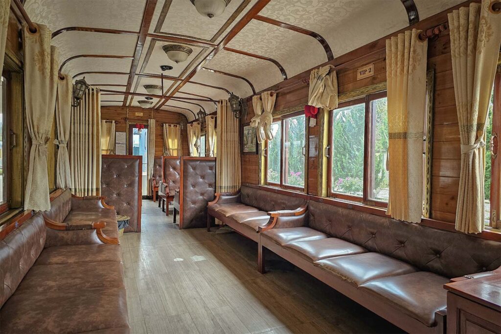 Interior of a vintage heritage train carriage in Dalat with brown tufted leather bench seats, warm wooden paneling, and cream curtains framing the windows