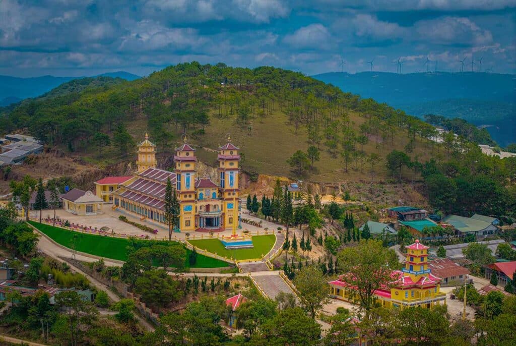 Aerial view of the Cao Dai temple in Dalat, a yellow building with three ornate towers set on a green lawn, backed by a pine-covered hill with wind turbines along the distant ridge
