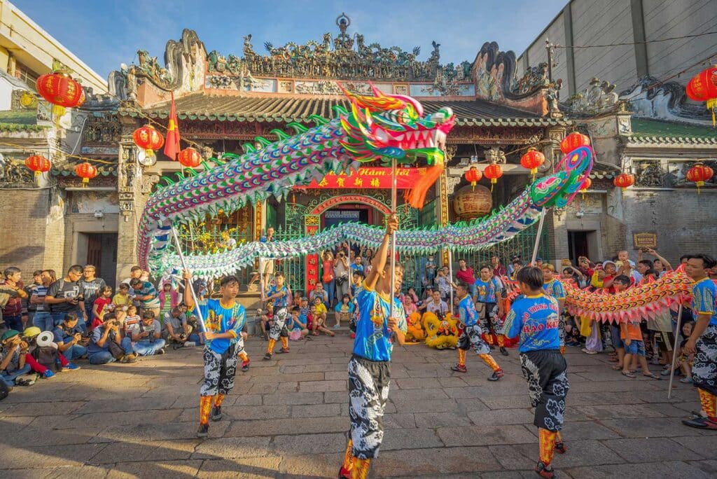 A colorful dragon dance performed by young men in blue costumes in front of Thien Hau Temple in Ho Chi Minh City, watched by a crowd, with red lanterns hanging above the ornate temple roof