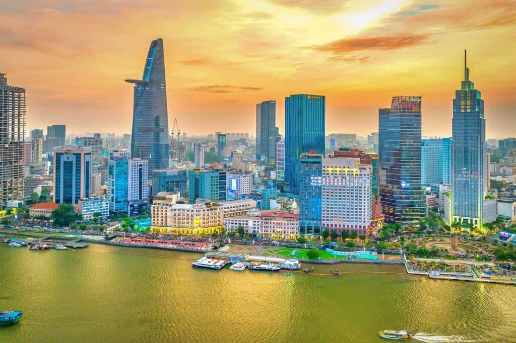 The Ho Chi Minh City skyline at sunset seen across the Saigon River, with the Bitexco tower and clusters of high-rise buildings glowing under an orange sky and boats along the waterfront
