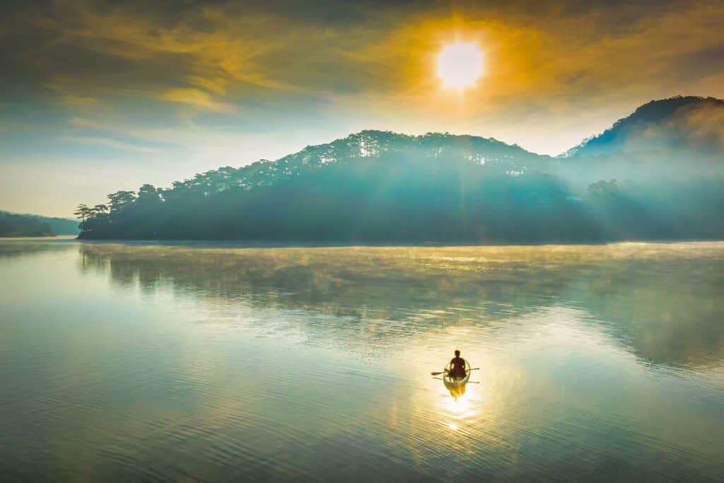 A person kayaking alone across the calm, misty water of Tuyen Lam Lake in Dalat at sunrise, with the sun rising over a forested hillside, a peaceful thing to do in Dalat