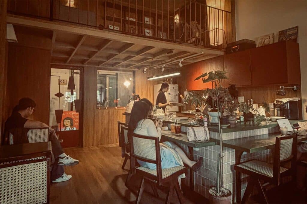 The warm wood-panelled interior of a quiet coffee shop in Ho Chi Minh City, with a tiled counter, mid-century rattan chairs, a mezzanine level, and a few people sitting with drinks