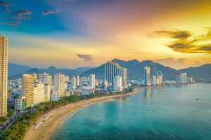 Aerial view of the Nha Trang city skyline at sunset, with high-rise hotels lining the curving bay, mountains behind, and a colorful orange and blue sky over the calm sea
