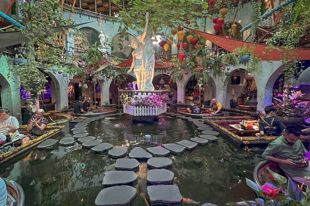 The plant-filled interior of a themed cafe in Ho Chi Minh City, with stepping stones across a koi pond, a central statue, hanging lanterns and greenery, and people seated in sunken booths around the water