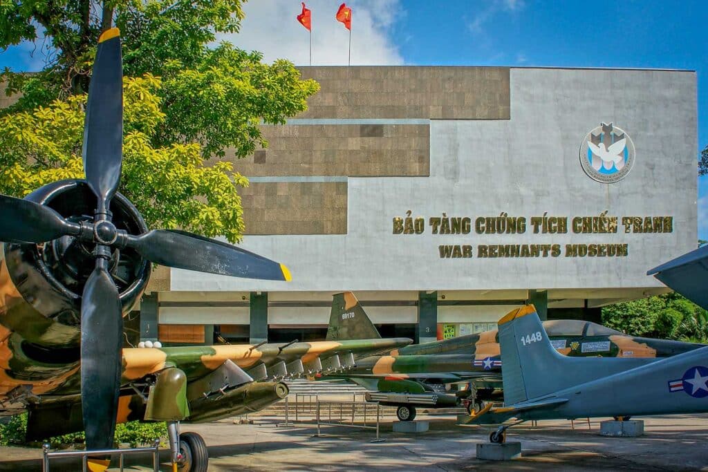 The outdoor display at the War Remnants Museum in Ho Chi Minh City, with camouflaged military aircraft and a large propeller plane parked in front of the grey museum building under Vietnamese flags