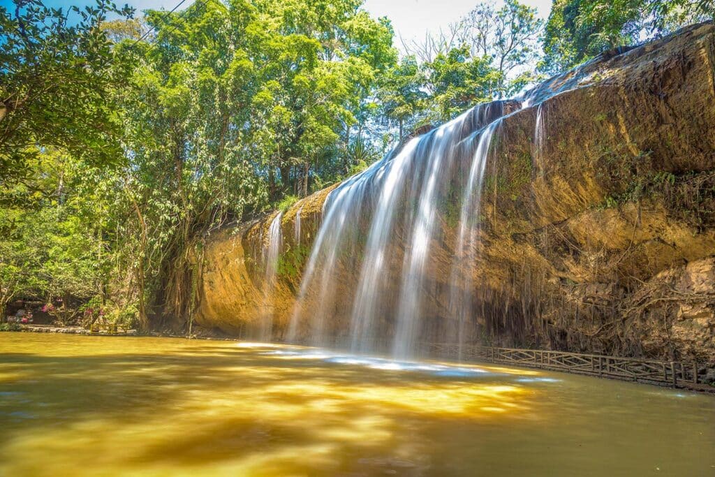Prenn Waterfall in Dalat spilling in wide thin curtains over a rock face into a golden pool below, framed by lush green forest and a wooden walkway on one side