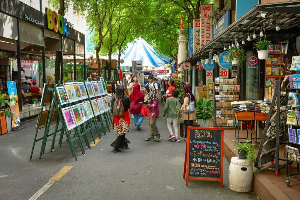 Nguyen Van Binh Book Street in Saigon, a tree-lined pedestrian lane with bookshops on both sides, framed prints displayed on stands, and visitors browsing among the greenery, one of the more relaxed things to do in Ho Chi Minh City