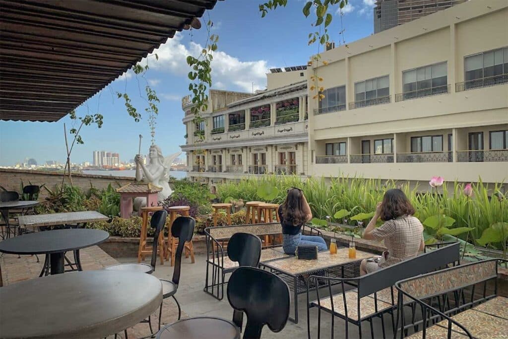 The rooftop terrace of a riverside cafe in Ho Chi Minh City, with metal tables, a white mermaid statue, lotus plants, and two women looking out over the Saigon River and old colonial buildings