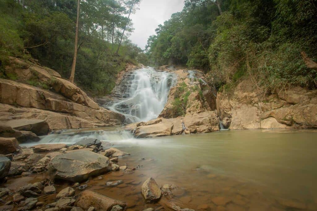 Tiger Waterfall near Dalat cascading over rocky steps into a calm green pool, surrounded by dense forest and large boulders in the foreground