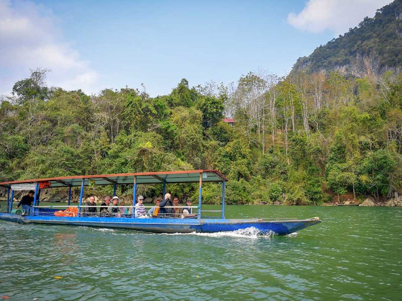 Travelers cruising on a traditional boat across the green waters of Ba Be Lake, with forested mountains in the background on a Ba Be Lake tour.