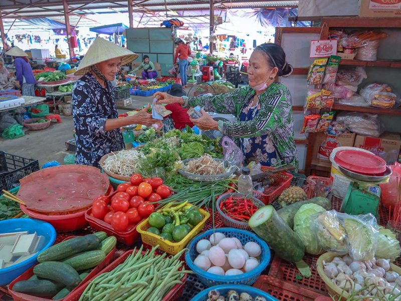 Woman buying fresh vegetables from a vendor at Ba Le Market in Hoi An, surrounded by tomatoes, cucumbers, herbs, and daily produce.