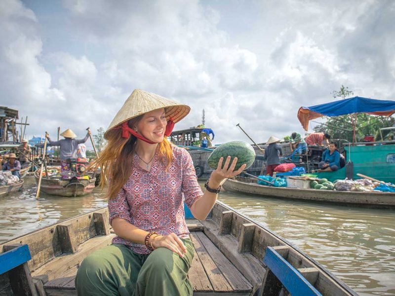 Traveler wearing a conical hat holding a watermelon on a boat ride at Cai Rang Floating Market in Can Tho.