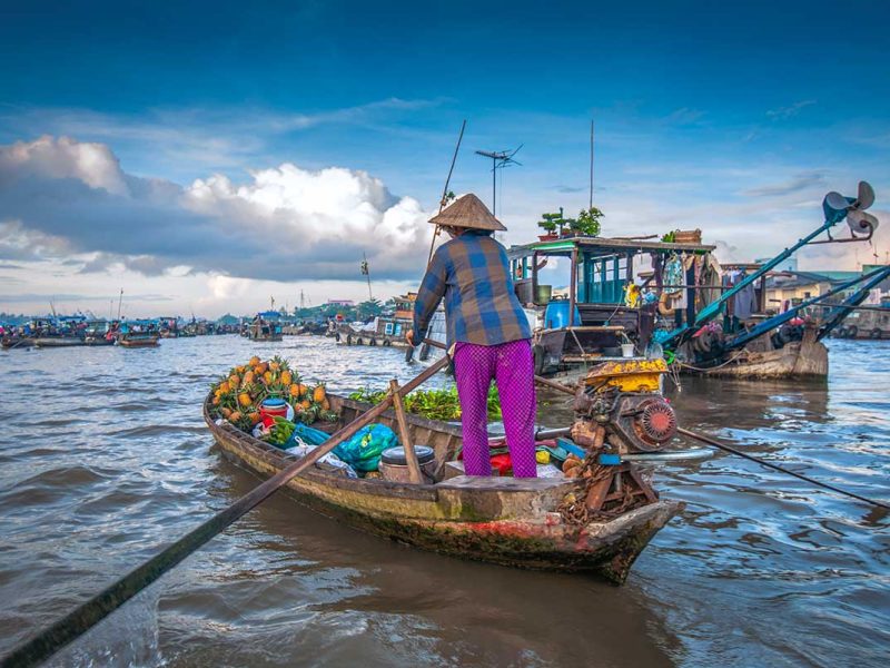 Sampan loaded with pineapples being rowed through Cai Rang Floating Market at sunrise, surrounded by larger cargo boats.