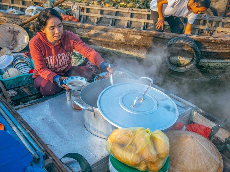 Local woman preparing steaming noodle soup from a floating kitchen boat at Cai Rang Floating Market, serving breakfast to visitors.