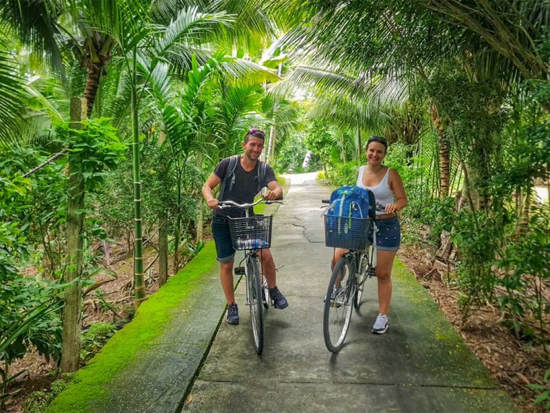Cycling tour through Can Tho village roads – Couple with bicycles on a narrow concrete path, surrounded by palm trees and lush greenery.