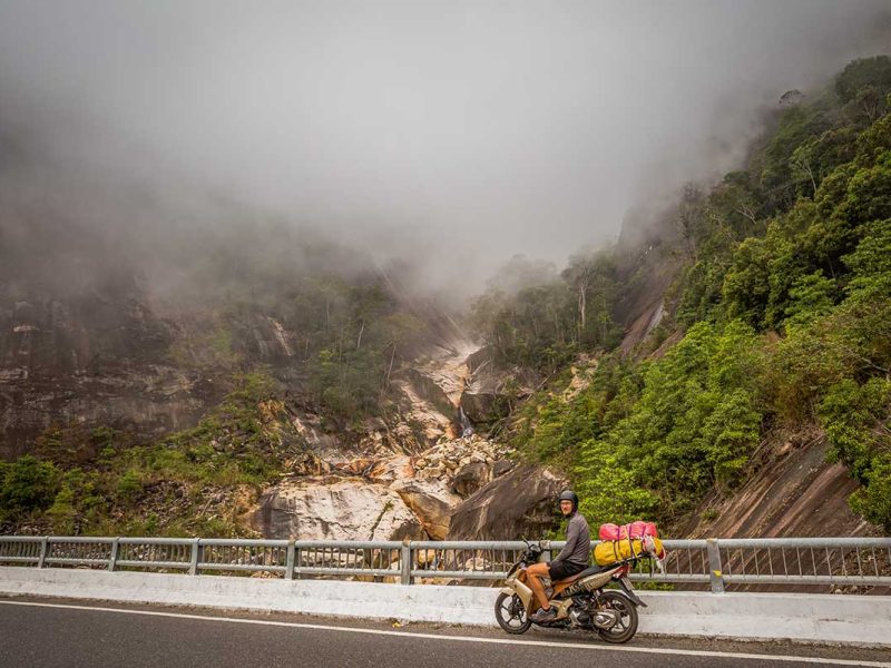 Motorbike rider stopping along the Dalat to Nha Trang route, with misty mountain scenery and waterfalls along the scenic pass