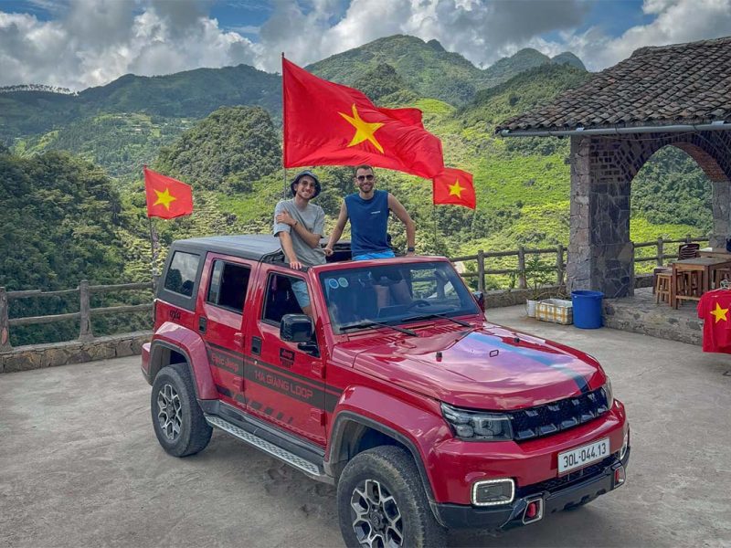 Local Vietnam guests on a Ha Giang Loop jeep tour at a mountain viewpoint, flying the Vietnamese flag during an actual tour stop, highlighting locally guided, real-world travel experiences in Ha Giang.