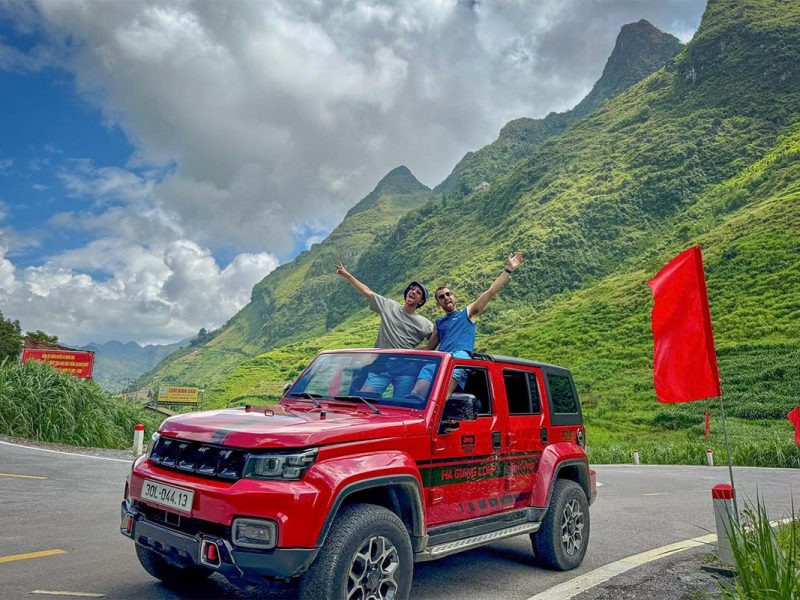 Guests standing through the open roof of a red jeep during a Ha Giang Loop jeep tour with Local Vietnam, captured on an actual mountain road stop surrounded by dramatic limestone scenery.