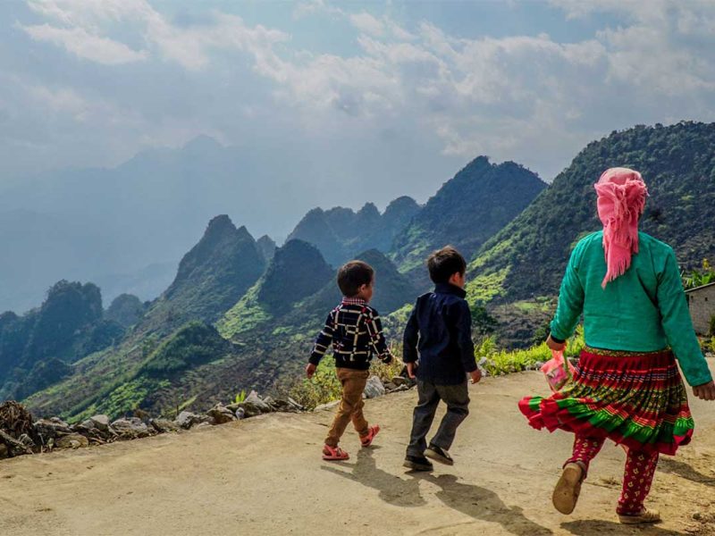 Ethnic minority woman and children walking along a mountain road on the Ha Giang Loop, with dramatic karst landscapes in the background