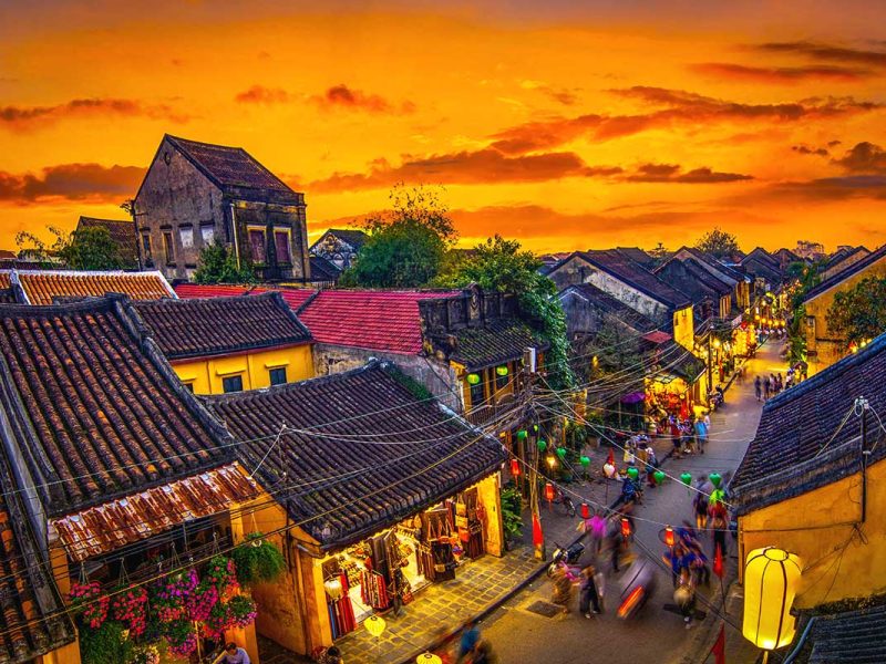 A view of the ancient town of Hoi An at sunset, creating a golden hue sky above the tiled roofs of the old houses.