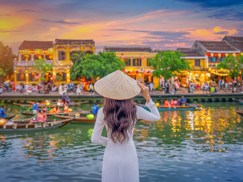 Woman wearing a traditional Vietnamese conical hat overlooking the lantern-lit river and yellow merchant houses of Hoi An Ancient Town at sunset.