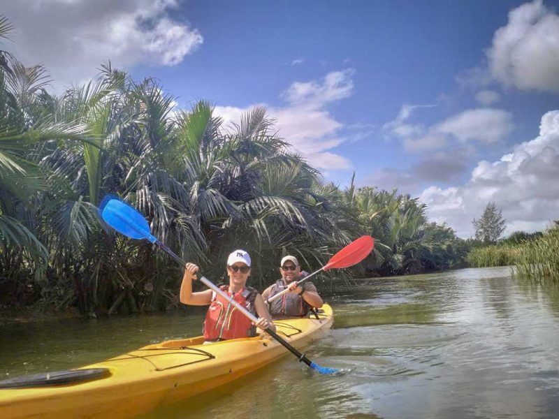 Two travelers kayaking through dense nipa palm mangroves under a bright sky during the Hoi An tour