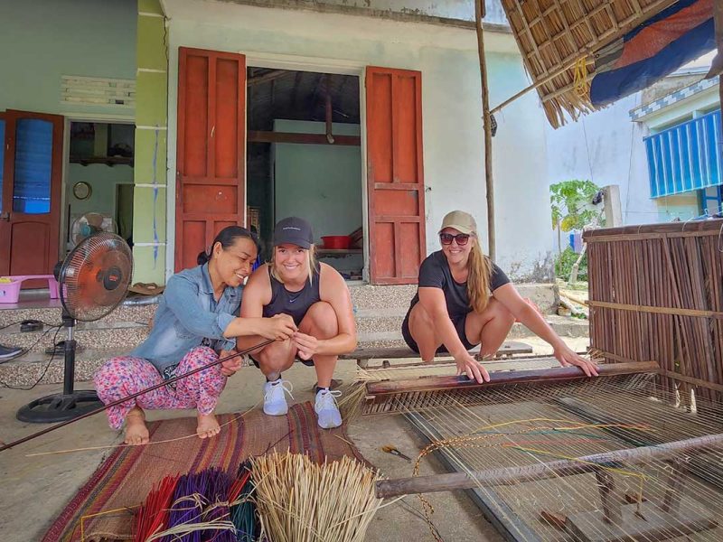 Two travelers learning traditional mat weaving with a local woman during the Hoi An countryside cycling tour