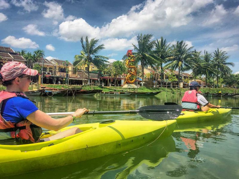 Two travelers kayaking on the Thu Bon River with palm trees and Hoi An Ancient Town scenery around them