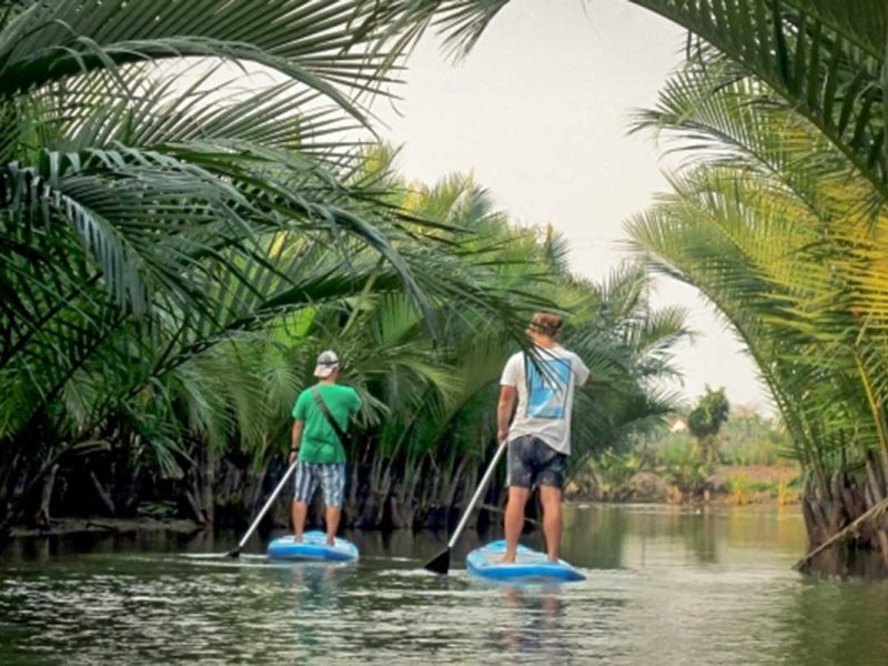 Two travelers stand-up paddling through a narrow tunnel of nipa palms on the Hoi An mangrove forest tour