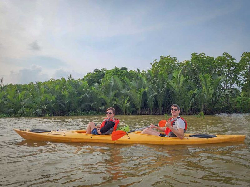 Two travelers kayaking across calm brown river water with mangrove palms in the background near Hoi An