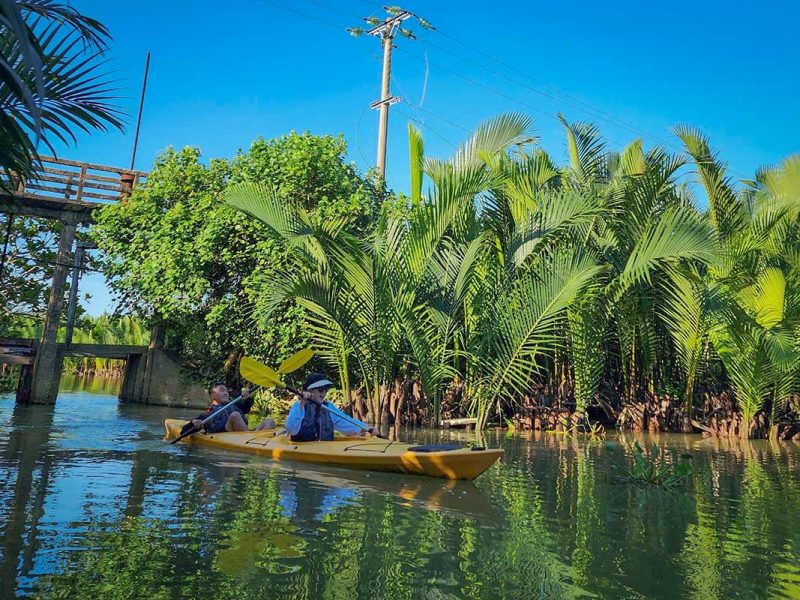 Travelers kayaking through a shaded mangrove passage surrounded by tall palm leaves in Hoi An