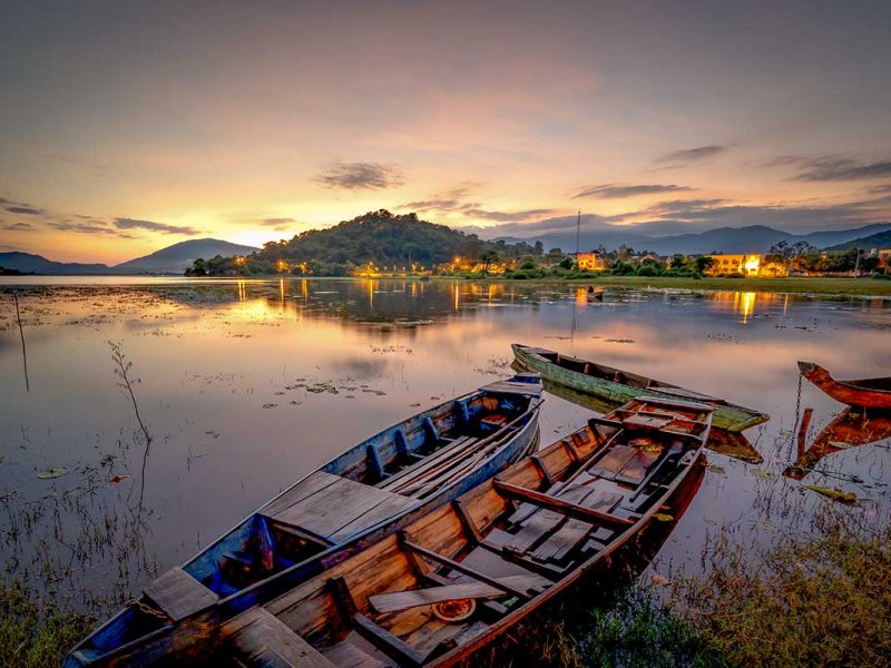 lak lake dak lak evening boats 1 Evening view of Lak Lake in Dak Lak with wooden boats moored in shallow water and village lights reflecting on the lake