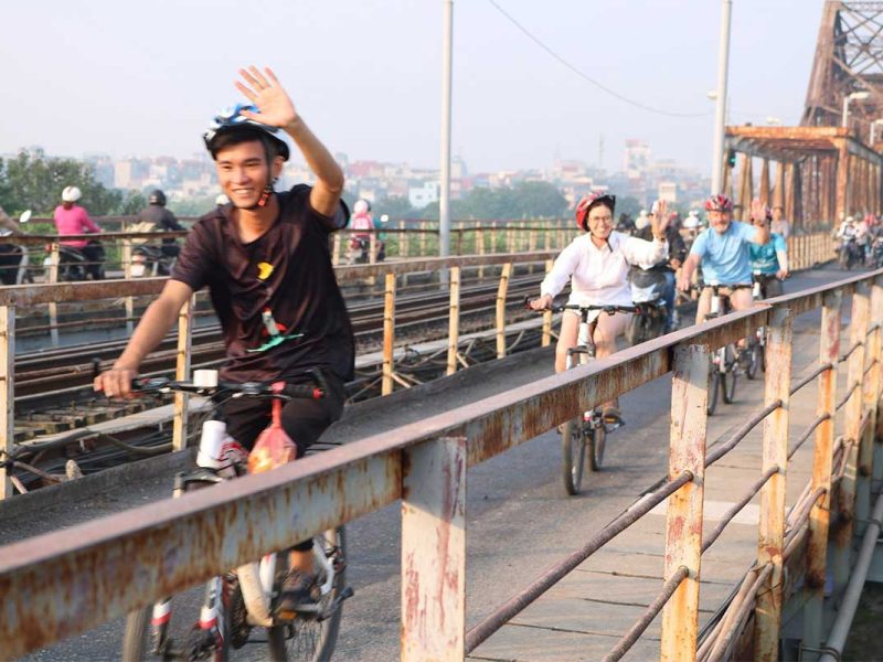 Local biking guide waving while leading tourists over Long Bien Bridge on a Hanoi bike tour