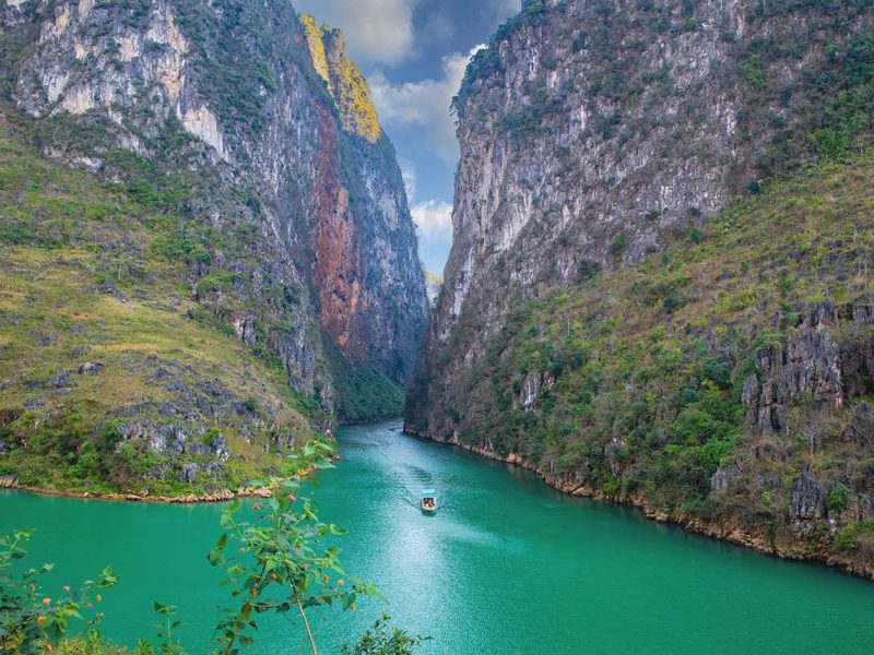 A distant view of a boat gliding through the emerald-green waters of Nho Que River, surrounded by the dramatic cliffs of Ma Pi Leng Pass.