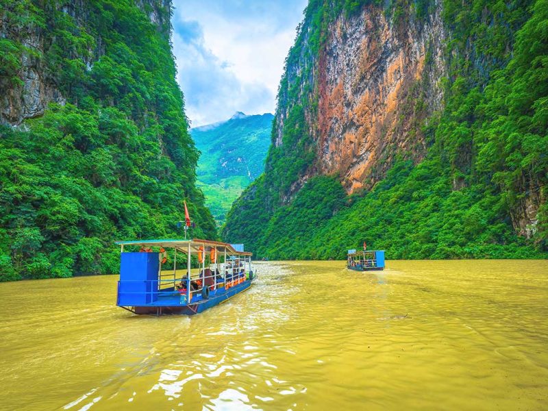 A tourist boat cruising along the brown waters of the Nho Que River, passing between the towering cliffs of Ma Pi Leng Pass in Ha Giang.