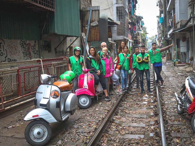 Group of travelers on Hanoi Train Street with Vespa motorbikes parked nearby during a motorbike tour