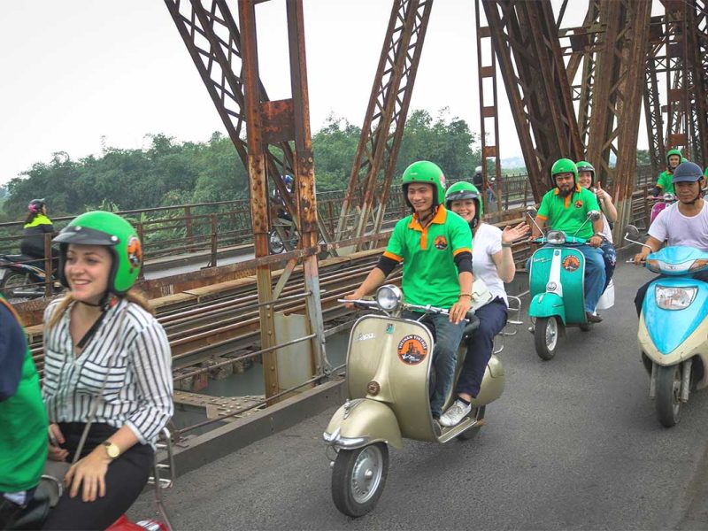 Hanoi motorbike tour with tourists riding over Long Bien Bridge on classic Vespa scooters