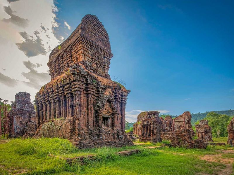 Weathered Cham brick tower under stabilization scaffolding at My Son Sanctuary, set in a grassy valley ringed by low mountains