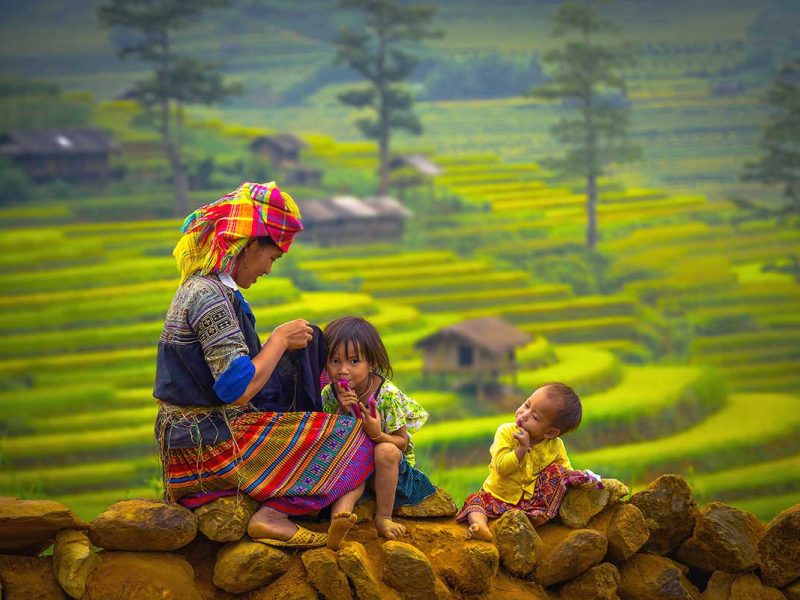 A colorful ethnic minority woman sitting on a stone wall with two children, with terraced rice fields of Sapa in the background.