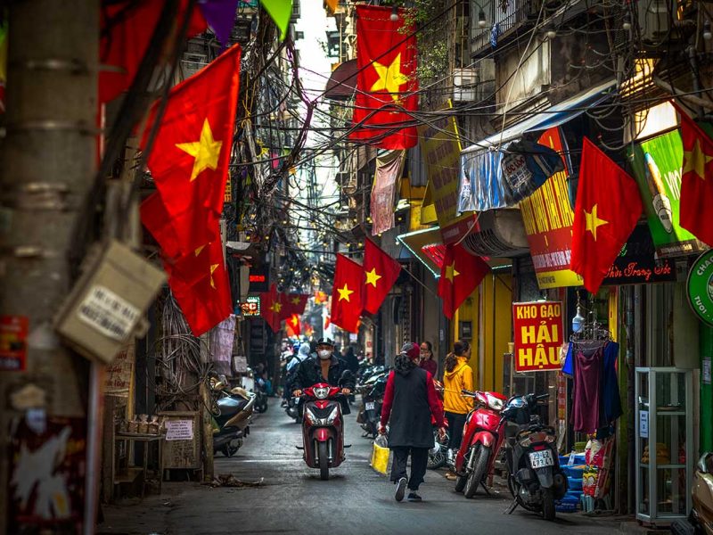 A bustling street scene in Hanoi, a vibrant city in Vietnam, with motorbikes zipping through narrow lanes, locals walking past colorful shophouses adorned with Vietnamese flags.