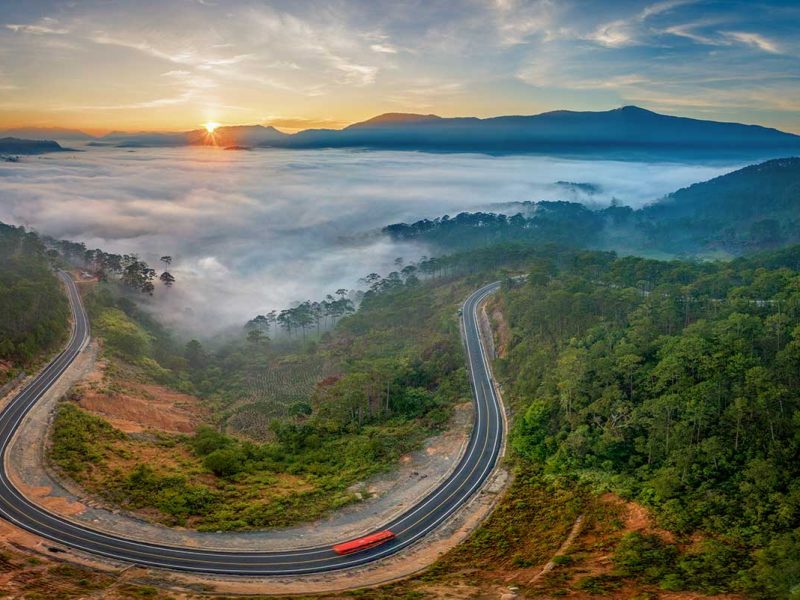 Aerial view of Khanh Le Pass at sunrise with clouds filling the valleys, showing winding mountain roads through forested hills