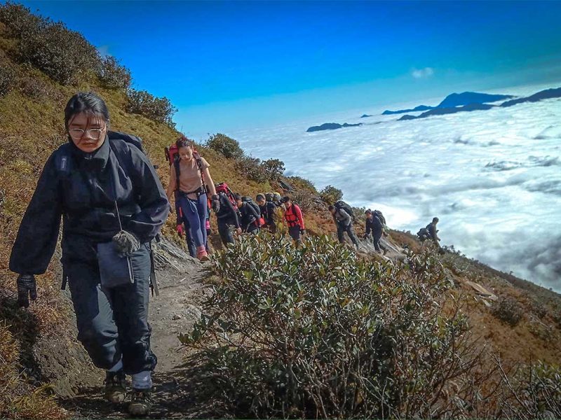 A group of tourists are trekking in Ta Xua Nature Reserve over a mountain path with in the background below them a sea of clouds