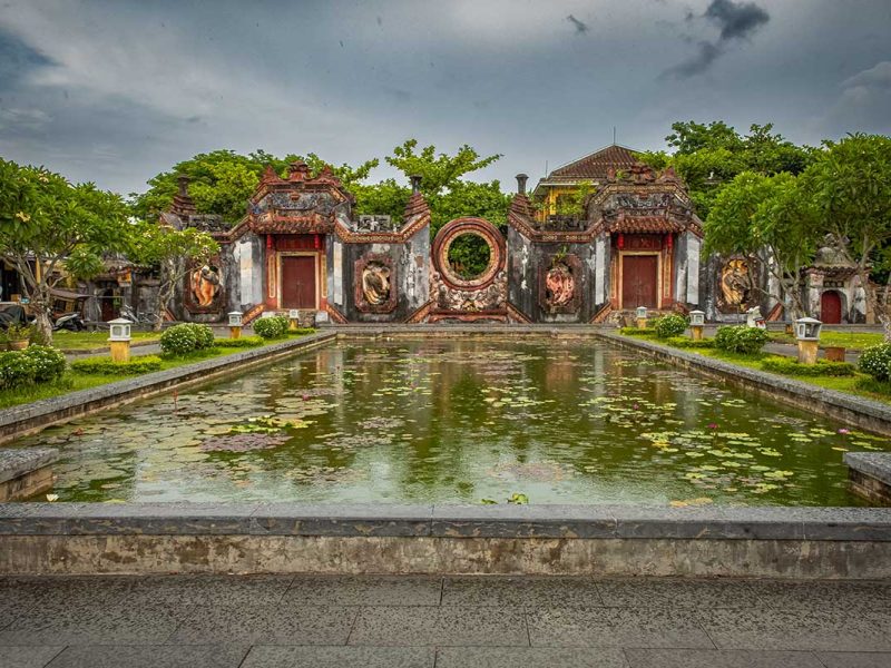 Tâm Ứng Bà Nữ Temple in Hoi An Ancient Town, showing the ancient courtyard, pond, and intricate old stone gates surrounded by greenery.