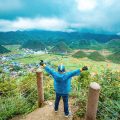 A man standing with arms raised in excitement at the viewpoint overlooking the Twin Mountains in Quan Ba.