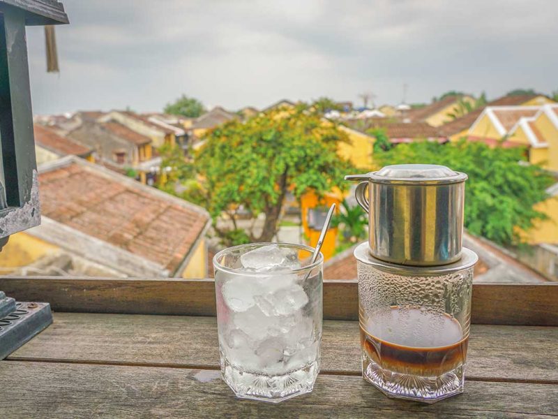 Traditional Vietnamese iced coffee served with a phin filter overlooking the yellow-tiled rooftops of Hoi An Ancient Town from a local café balcony.